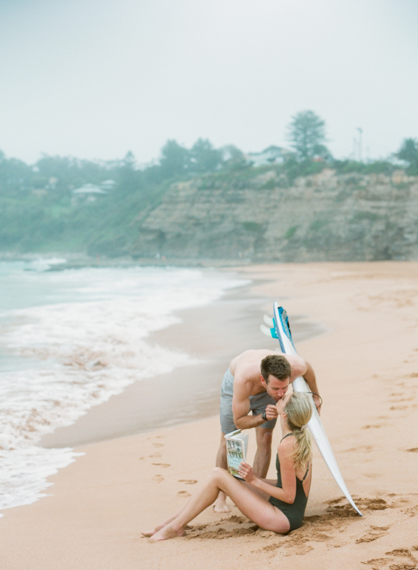 Fiance holding surf board kissing bride to be at Avalon beach in sydney for an engagement shoot with Amelia Soegijono who is a fine art film photographer